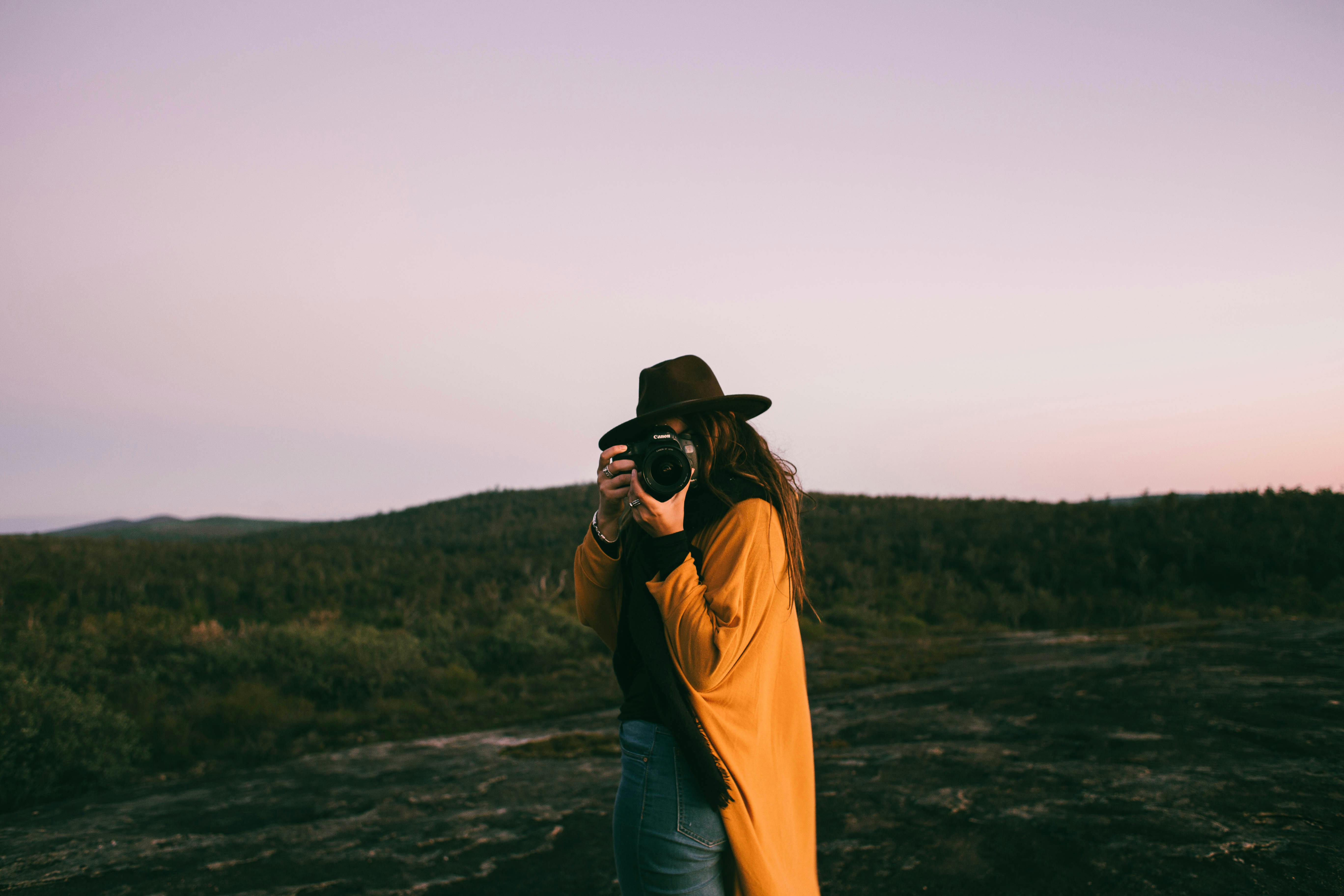 Es una chica con sombrero en un paisaje árido sacando una fotografía dirigiendo su cuerpo hacia la cámara.